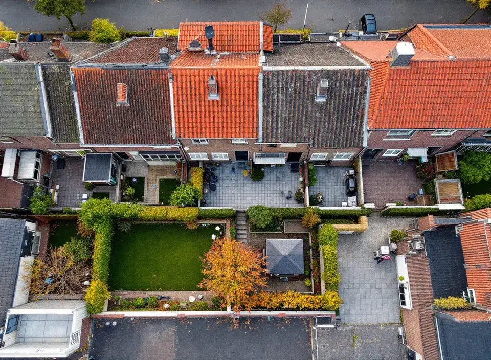 Smalle achtertuinen achter een rij bakstenen rijtjeshuizen, van bovenaf gezien, met gazon, terrassen en schuurtjes
